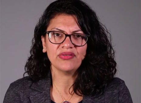 A person with curly dark hair and glasses, wearing a dark blazer and a necklace, against a plain background.