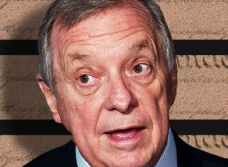 An older man with gray hair wearing a suit and tie, standing in front of a background with text and horizontal lines.