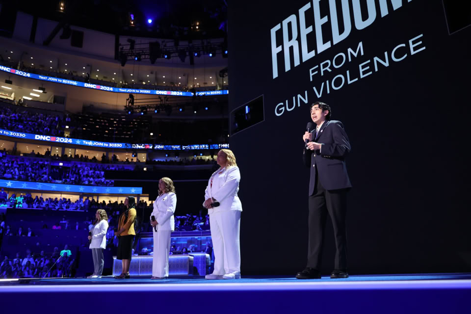 Five people stand on a stage with a large screen behind them displaying the words "Freedom from Gun Violence." The venue is filled with an audience, and the lighting is predominantly blue.