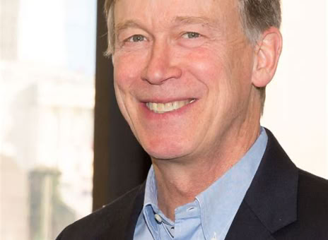 A man in a dark suit and light blue shirt smiling, standing indoors with a window in the background.