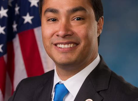 A man in a suit with a blue tie, standing in front of an American flag.