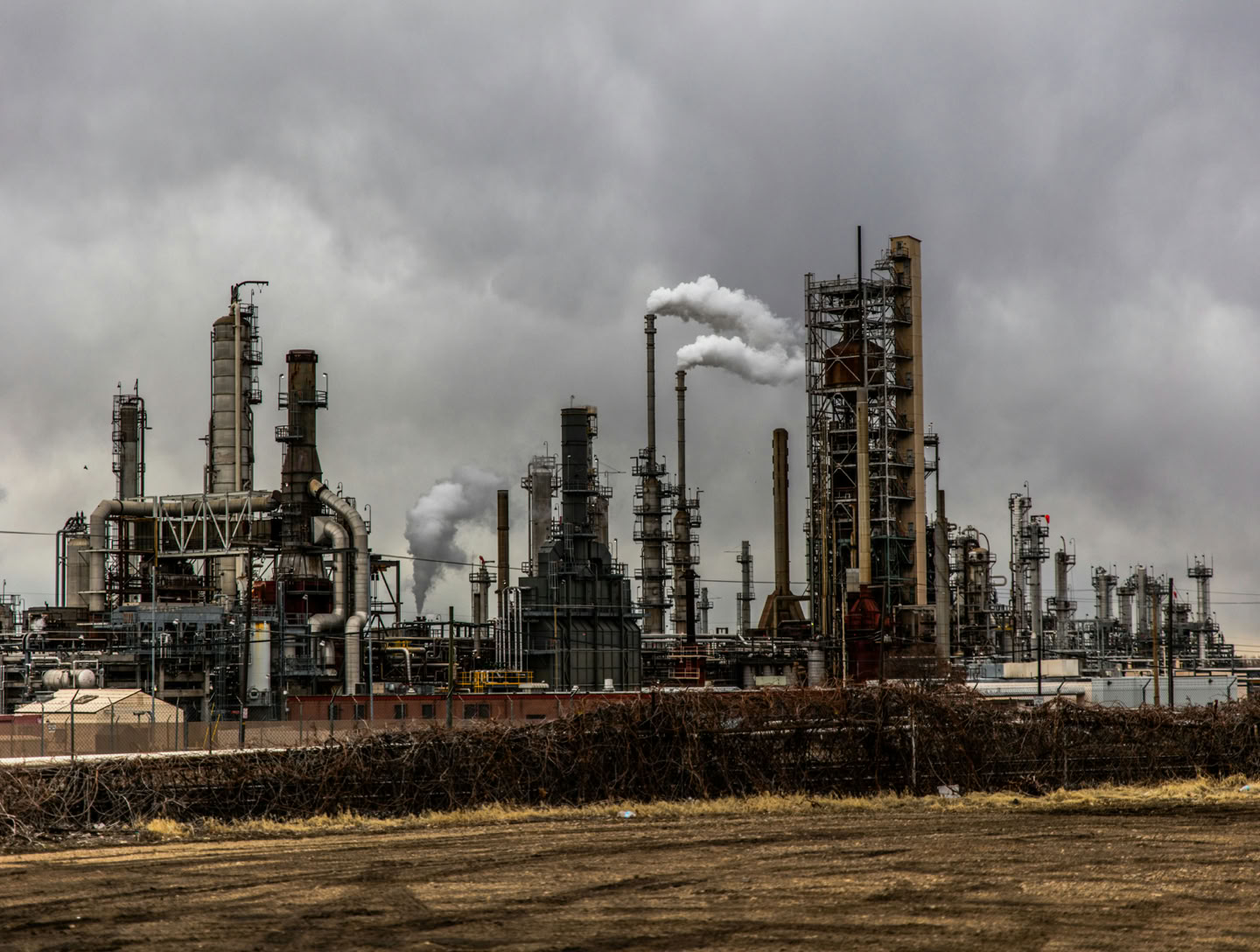 Industrial refinery complex with multiple smokestacks emitting smoke under a cloudy sky.