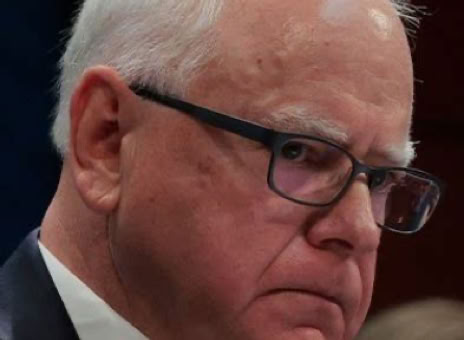 Tim Walz with white hair and glasses, wearing a suit and tie, looking serious.