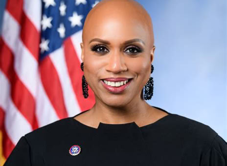 A person with a shaved head wearing a black outfit and earrings, standing in front of an American flag.