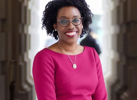 A person with curly hair and glasses, wearing a pink dress and a necklace, stands smiling in an outdoor corridor with columns.