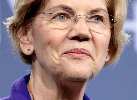 A person with short, light-colored hair and glasses, wearing a purple blazer, against a blurred background.