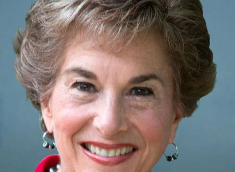 A person with short, light brown hair, wearing a red top and pearl earrings, smiling against a gray background.