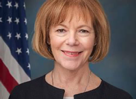 A woman with short, light brown hair wearing a black blazer, standing in front of an American flag and a blue background.