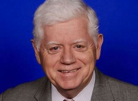An older man with white hair wearing a gray suit, white shirt, and red tie, against a blue background.