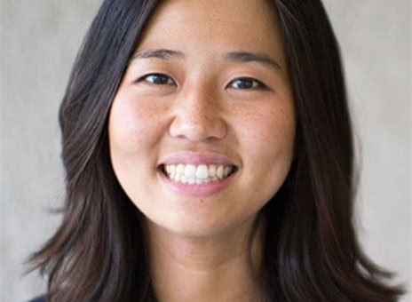 A person with shoulder-length dark hair smiling, wearing a dark blazer, against a neutral background.