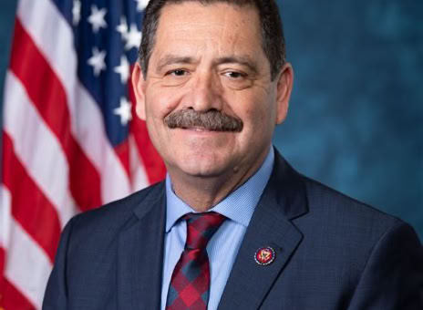 A man in a suit and tie is standing in front of an American flag with a blue background.