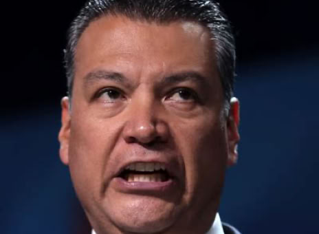 A person speaking with an expressive face, wearing a suit and tie, against a dark background.