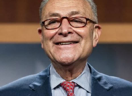 A man wearing glasses, a suit, and a red tie, smiling in front of a blurred background.