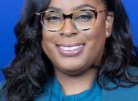 A person with long dark hair and glasses, wearing a teal outfit with a pin, against a blue background.