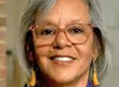 An older adult with gray hair, wearing glasses, a purple blazer, and yellow earrings, smiling.