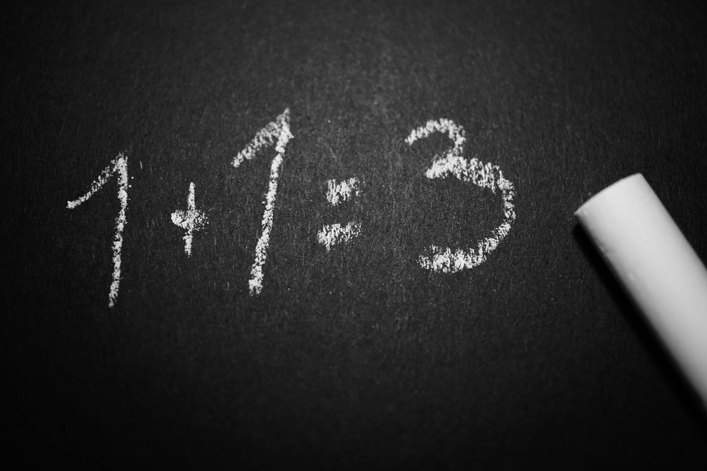 Chalkboard with the equation "1 + 1 = 3" written in white chalk, with a piece of chalk lying nearby.