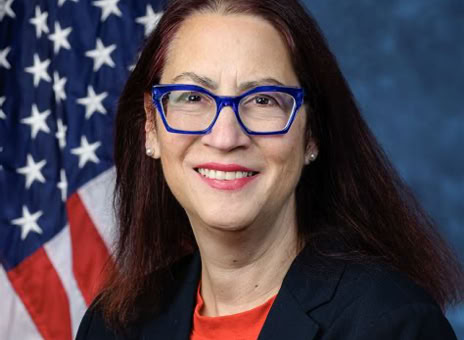 A person with long dark hair and blue glasses, wearing a black blazer and red shirt, stands in front of an American flag with a blue background.