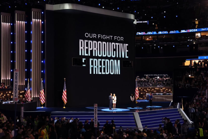 A person stands on a stage with a large screen displaying the text "Our Fight for Reproductive Freedom." The stage is surrounded by American flags and an audience is visible.
