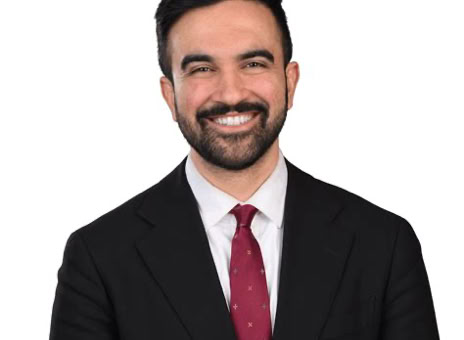 A man with dark hair and a beard is smiling, wearing a black suit, white shirt, and red tie, against a white background.