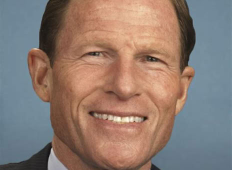A man in a suit and tie smiling against a plain blue background.