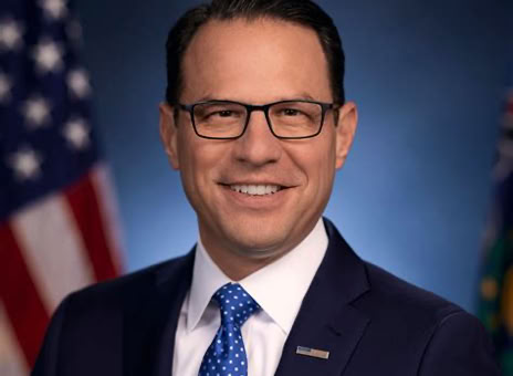 A man in a suit and tie, wearing glasses, stands in front of a blurred background with flags.